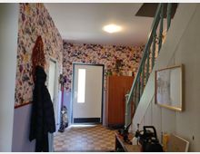 Flur eines Wohnhauses mit geblümter Tapete, einer hellen Eingangstür mit Glaseinsatz und einer Holztreppe mit mintgrün gestrichenen Geländern. Im Raum befindet sich ein Holzschrank, verschiedene Gegenstände auf einer Werkbank und ein gerahmtes Bild an der Wand.