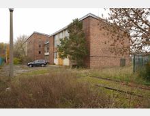 Backsteinbau mit flachem Dach in einem verlassenen, überwucherten Gelände mit einem geparkten schwarzen SUV, Eisenbahnschienen und Laternenmasten. Im Vordergrund befinden sich Gras, Büsche und ein Zaun rechts am Bildrand.