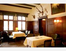 Ein traditionelles Restaurant mit holzvertäfelten Wänden, dekoriert mit Hirschgeweihen und einem Gemälde. Die Einrichtung umfasst mit weißen Tischdecken gedeckte Tische, grüne gepolsterte Stühle und große Fenster mit Vorhängen, die weiches Tageslicht hereinlassen.