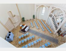 Blick in den Innenraum einer kleinen Kapelle mit hellen Wänden und Ziegelboden. Der Raum ist für eine Veranstaltung vorbereitet, mit in Reihen angeordneten Stühlen mit blauen Sitzkissen, einem dekorierten Tisch vorne sowie einer Kanzel und einem Treppenaufgang auf der rechten Seite.