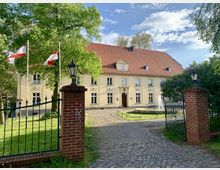 Gelbes, zweistöckiges Herrenhaus mit rotem Ziegeldach, umgeben von Bäumen, mit einem gepflasterten Weg und einem Brunnen im Vorgarten. Im Vordergrund ein Eingangstor mit Ziegelpfosten und schwarzem Metallzaun, auf einer Seite wehen zwei Flaggen.