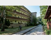 Mehrstöckiges Wohngebäude mit Balkonen, gelben Fassaden und braunen Akzenten, umgeben von Bäumen und Grünflächen. Ein gepflasterter Weg und ein schmaler Fußweg führen an der Fassade vorbei, im Vordergrund ein angelehntes Fahrrad.