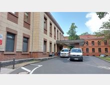 Außenansicht eines Krankenhausgebäudes mit sandfarbener und roter Backsteinfassade, mehreren Fenstern und einem überdachten Eingangsbereich. Zwei Krankenwagen stehen vor dem Eingang, umgeben von gepflasterten Wegen und Bäumen im Hintergrund.