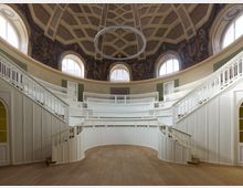 Innenansicht eines historischen Rundbaus mit Holzvertäfelung und geschwungenen weißen Balustraden. Die Kuppeldecke ist kunstvoll gestaltet, mit geometrischen Mustern und Wandmalereien darunter; große Fenster lassen Tageslicht in den Raum strömen.