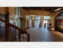 Innenansicht eines Gebäudeeingangs mit Fliesenwänden, Holztüren mit Glaselementen und einem gefliesten Boden. Im Vordergrund ist ein Treppengeländer zu sehen, während im Hintergrund mehrere Personen durch geöffnete Türen auf eine Straße gehen.