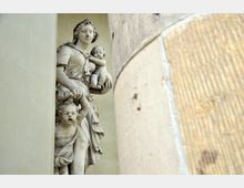 Steinskulptur einer Frau mit zwei Kindern, teilweise verdeckt von einer massiven Steinsäule. Die Figuren sind detailliert gearbeitet und in klassischem Stil gehalten, vor einem beigen, glatten Hintergrund.