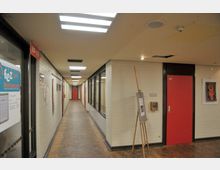 Ein langer Flur in einem Innenbereich mit cremefarbenen Backsteinwänden, roten Türen und einer Holzparkett-Bodenfläche. Auf der rechten Seite steht eine Staffelei mit einem Plakat, und links befinden sich Glasfenster, die in einen angrenzenden Raum blicken.