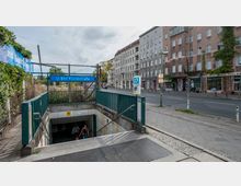 U-Bahnhof-Eingang zur Station "Yorckstraße" mit blauem Schild und Geländern, umgeben von Straßenpflaster, einer angrenzenden Straße und Wohngebäuden im Hintergrund. Im Vordergrund führen Treppen ins unterirdische Stationsgebäude.