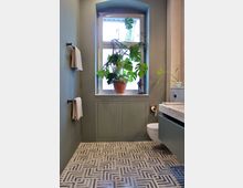 Modernes Badezimmer mit mattgrünen Wänden und einem bodenstehenden Fenster mit Topfpflanze (Monstera) auf dem Sims. Der Boden hat geometrische Fliesenmuster, und neben dem schwebenden Waschtisch befindet sich ein wandmontiertes WC.