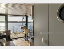 Innenraum eines modernen Hausbootes mit großen Fenstern, die den Blick auf das Wasser und das Ufer im Hintergrund freigeben. Im Vordergrund sind ein Steuerrad, ein Sitzplatz und ein Teil der kleinen Küche sichtbar, während viel Licht den Raum erhellt.