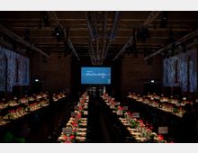 Großer Veranstaltungsraum mit Backsteinwänden, langen, festlich gedeckten Tischen und Blumenarrangements; eine Bühne mit einem Bildschirm im Hintergrund zeigt den Schriftzug „Berliner Meisterköche 2019“.