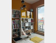 Ein kleines Wohnzimmer oder Arbeitszimmer mit gelb-orange gestrichenen Wänden, einem Regal mit Büchern und Aufbewahrungsboxen, einem modernen Stuhl und einer großen Fensterfront, die Tageslicht hereinlässt. Im Hintergrund sind ein Globus sowie eine moderne Hängelampe mit runden Elementen sichtbar.
