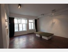 Ein leerer Raum mit weißen Wänden, einem dunkelroten Betonboden und großen Fenstern mit schwarzen Vorhängen. In der Mitte des Raumes steht eine niedrige Sitzbank mit grün gepolsterten Sitzflächen.