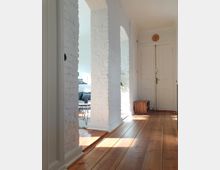 Flur eines Wohnhauses mit Holzdielenboden, weiß gestrichenen Backsteinbögen und einer doppelflügeligen weißen Tür im Hintergrund. Durch die Bögen ist ein Wohnbereich mit einem Sofa und einem kleinen runden Couchtisch sichtbar.