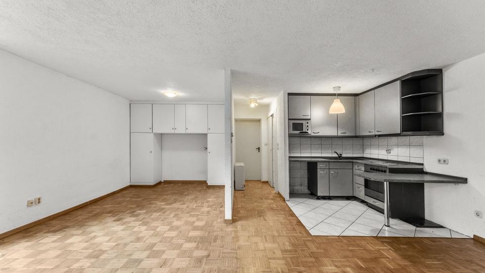 Offener Wohnraum mit Parkettboden und weißer Decke; links sind Einbauschränke an der Wand, mittig führt ein Flur zu Türen. Rechts befindet sich eine L-förmige Küchenzeile mit grauen Schränken, Fliesenrückwand und -boden, Spüle und Herd.