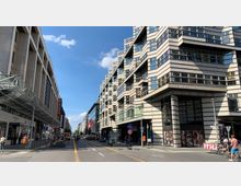 Städtische Straße mit modernen Gebäuden, darunter ein mehrstöckiges Gebäude mit markanter Fassade aus Glas und hellen Steinplatten. Links sind Geschäfte und ein überdachter Gehweg zu sehen, während die Straße durch gelbe Markierungen für Fahrradwege hervorgehoben ist.