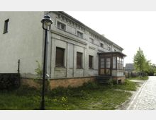 Ein schlichtes, zweistöckiges Wohngebäude mit verputzter Fassade und geschlossenen Rollläden. Vor dem Haus befindet sich eine Laterne und eine kleine Grünfläche, während rechts eine gepflasterte Straße mit Bäumen verläuft.