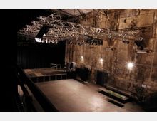 Ein leerer Veranstaltungsraum mit moderner Lichttechnik, einer kleinen Bühne mit schwarzem Vorhang im Hintergrund und einer hohen, verwitterten Betonwand mit Graffiti. An der Decke befindet sich ein Gerüst mit Scheinwerfern und Lautsprechern.