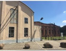 Zwei historische Gebäudefassaden, links ein beige verputzter Bau mit Metallfenstern und rostigem Schiebetor, rechts ein roter Backsteinbau mit hohen Rundbogenfenstern; im Vordergrund ein gepflasterter Weg mit großen Steinen und einer Straßenlaterne.