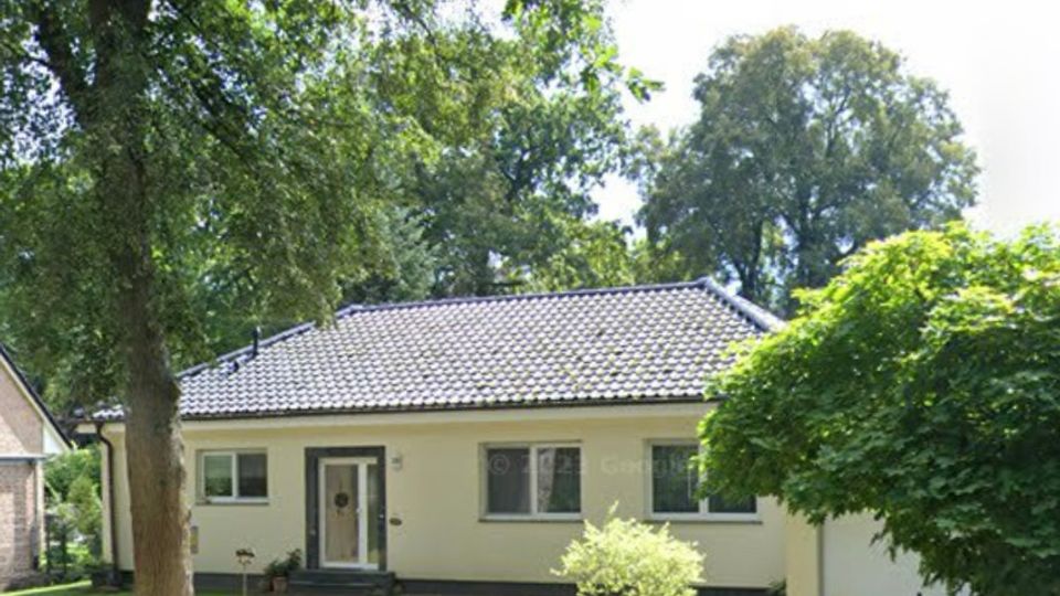 Einfamilienhaus im Bungalowstil mit hellem Putz und dunklem Ziegeldach, davor ein kleiner Vorgarten mit Rasen, Sträuchern, Steinen und großen Bäumen. Rechts führt eine gepflasterte Einfahrt zur angebauten Garage.