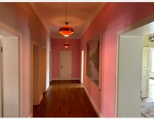 Ein schmaler Flur mit rosa Wänden, Holzboden und einer geschlossenen weißen Tür am Ende. An der Decke hängen mehrere orangefarbene Lampen, an der rechten Wand ist ein gerahmtes Bild zu sehen, und es gibt offene Türen, die zu angrenzenden Räumen führen.