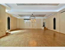 Ein leerer Raum mit hellen Backsteinwänden, einem Holzboden und einer großen modernen Hängeleuchte in der Mitte. An der Decke befinden sich verzierte Deckenelemente, und an der rechten Seite führen Stufen zu einem erhöhten Bereich.