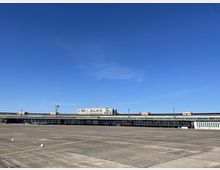 Außenansicht des ehemaligen Flughafens Berlin-Tempelhof. Zu sehen sind das weitläufige Flughafenvorfeld mit Betonflächen und das langgestreckte, denkmalgeschützte Terminalgebäude unter einem klaren blauen Himmel.