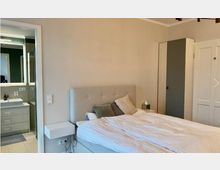 Schlafzimmer mit einem grauen Polsterbett, weißer Bettwäsche und mehreren Kissen. Links befindet sich der Zugang zu einem angrenzenden Badezimmer mit modernem Waschbecken und Spiegel, rechts ein schmaler Kleiderschrank und eine weiße Tür.