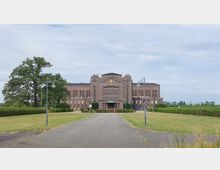 Backsteingebäude im symmetrischen Stil mit hohen Fenstern und mittigem Eingangsbereich, davor ein gepflasterter Weg, flankiert von Rasenflächen und Bäumen; umgeben von einer ländlichen Umgebung unter bewölktem Himmel.