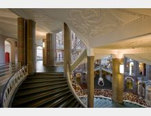 Großzügiger Treppenbereich in einem historischen Gebäude mit geschwungenen Treppen, dekorativen, weißen Geländern und verzierten Decken mit Stuck. Die Architektur wird von roten und sandfarbenen Säulen sowie weiten Bögen geprägt, die Tageslicht durch große Fenster hereinlassen.