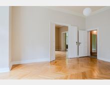 Heller, leerer Wohnraum mit hohen Decken, weiß gestrichenen Wänden und Fischgrätparkett aus Holz. Mehrflügelige weiße Doppeltüren führen in angrenzende Räume und einen Flur; an der Decke hängt eine runde Pendelleuchte.