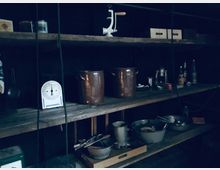 Dunkle Vorratskammer mit Holzregalen, auf denen kupferne und metallene Töpfe, eine Waage, Flaschen, sowie Küchenutensilien wie Schüsseln und Kellen stehen. Die Umgebung wirkt rustikal und funktional.