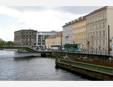 Stadtansicht mit einer Brücke über einen Fluss, umgeben von historischen und modernen Gebäuden. Im Vordergrund ein Wasserkanal mit einem Frachtkahn, auf der Brücke und Straße darüber fahren Busse und Fahrräder.