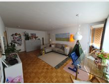 Wohnzimmer mit Parkettboden, einer beige Couch, einem Teppich und einer Kinderspielecke mit grünem Spielzelt. Rechts stehen ein Esstisch mit Glasplatte und Stühlen sowie eine große Fensterfront mit Zugang zu einem Balkon.