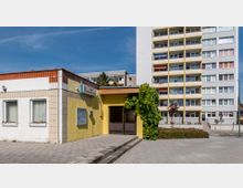 Einstöckiges gelb-weißes Gebäude mit flachem Dach und dem Schriftzug "Zum Schwarzen Peter" über dem Eingang, umgeben von Grünpflanzen. Im Hintergrund steht ein mehrstöckiges Wohnhaus mit Balkonen und bunt gestalteten Fassaden.