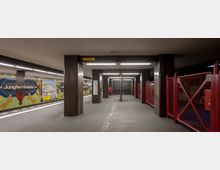 Ein U-Bahnhof mit breiter Bahnsteigfläche, grauem Boden und dunklen Säulen. Die Wände sind mit bunten Mosaiken gestaltet, die den Schriftzug „Jungfernheide“ enthalten, und ein gelbes Schild weist den Ausgang aus.