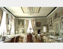 Ein eleganter Speisesaal im Vintage-Stil mit pastellfarbenen Wänden, verzierten Tapeten und Dekorationen wie Kronleuchtern, Gemälden an den Wänden und weißen Tischdecken. Der Raum ist mit antiken Möbeln, Kerzenhaltern und dezenten Blumenarrangements ausgestattet.