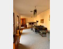 Wohnzimmer mit Teppichboden, einer grauen Couch und einem kleinen Couchtisch. Im Hintergrund befinden sich eine Küche mit Barhockern und eine hölzerne Bücherregalwand, ergänzt durch helle Wände und hängende Pflanzen.