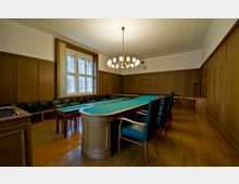 Ein Sitzungssaal mit Holzwänden, einem langen Tisch mit grüner Tischoberfläche und gepolsterten Stühlen. Ein großes Fenster mit weißen Rahmen und ein Kronleuchter bilden zentrale Elemente; der Boden ist aus Holz.