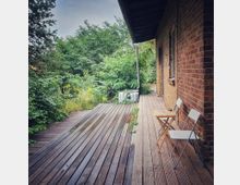 Holzterrasse neben einem Backsteingebäude, mit zwei Klappstühlen und einem kleinen Tisch; umgeben von dichter, grüner Vegetation und Bäumen. Ein Abflussrohr hängt von der Dachkante herab, und die Terrasse zeigt Spuren von Regen.