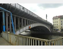 Eisenbahnbrücke über einem Kanal mit stählerner Tragwerkskonstruktion und hellem Geländer im Vordergrund. Im Hintergrund stehen Wohngebäude und ein Café mit Außensitzplätzen.