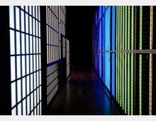 Ein schmaler, dunkler Korridor mit beleuchteten Wänden aus transparenten Paneelen auf beiden Seiten; links in Weiß und Blau, rechts in Blau und Grün. Der Boden ist dunkel reflektierend und der Hintergrund ist in Schatten getaucht.