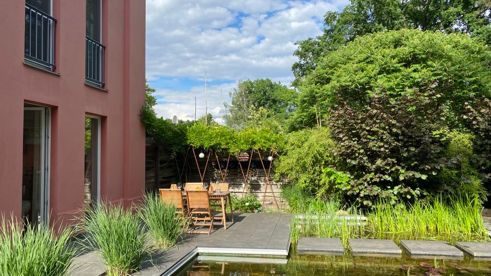 Moderner Gartenhof neben einem rosafarbenen Gebäude mit großer Terrassenfläche, Holztisch und Stühlen unter einer begrünten Pergola. Im Vordergrund liegt ein rechteckiger Naturteich mit Seerosen, Schilfpflanzen und Trittplatten, umgeben von dichtem Baumbestand.