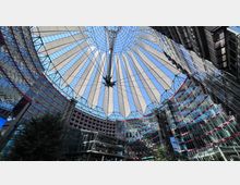 Modernes architektonisches Areal mit einer großen gläsernen Struktur, überdacht von einem auffälligen, zeltähnlichen Dach aus weißen Streben und Metallträgern, das von einer zentralen Spitze ausstrahlt. Umgeben von gläsernen Fassaden mehrerer Gebäude mit roten Akzenten, die einen offenen Hof mit Bäumen und Sitzgelegenheiten einschließen.