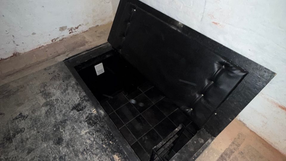 Kellerraum mit weiß verputzten Wänden und Steinboden; im Boden befindet sich eine rechteckige, geöffnete Bodenluke mit schwarzer Klappe, die in einen dunklen, gefliesten Schacht führt. An der Wand verlaufen sichtbare Kabel und Leitungen.