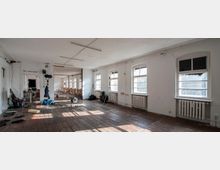 Großer, heller Raum mit weißen Wänden, Holzfußboden und mehreren Fenstern mit Heizkörpern darunter. Der Raum wirkt unfertig und ist mit Stühlen, Kameraständern, Kabeln und anderem technischen Equipment teils befüllt.