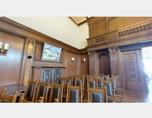 Holzvertäfelter Raum mit Stuhlreihen, verziert mit dunklen Lederpolstern und Nieten. Die Wände sind mit dekorativen Holzschnitzereien und einem Landschaftsbild geschmückt, außerdem sind kleine Wandlampen und ein kunstvoll gestalteter Kamin sichtbar.