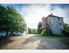 Historisches Gebäude mit Fachwerkelementen und Bruchsteinmauerwerk, daneben ein weißes Haus und ein gepflasterter Hof mit Bäumen und Grünflächen im Hintergrund. Der Himmel ist leicht bewölkt, und die Szene ist von natürlichem Tageslicht beleuchtet.