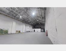 Großer, leerer Innenraum mit hohen, weiß getünchten Wänden, einem offenen Dachgebälk und Hängelampen. An den Wänden stehen Gemälde und Gegenstände; im Hintergrund ist ein kleiner, separater, weißer Raum erkennbar.