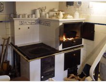 Historischer Küchenofen mit weiß gefliester Verkleidung und dekorativen blauen Kacheln mit der Aufschrift „Sich regen bringt Segen“. Der Ofen hat eine geöffnete Klappe mit sichtbarem Feuer und Küchenutensilien auf der Ablage.